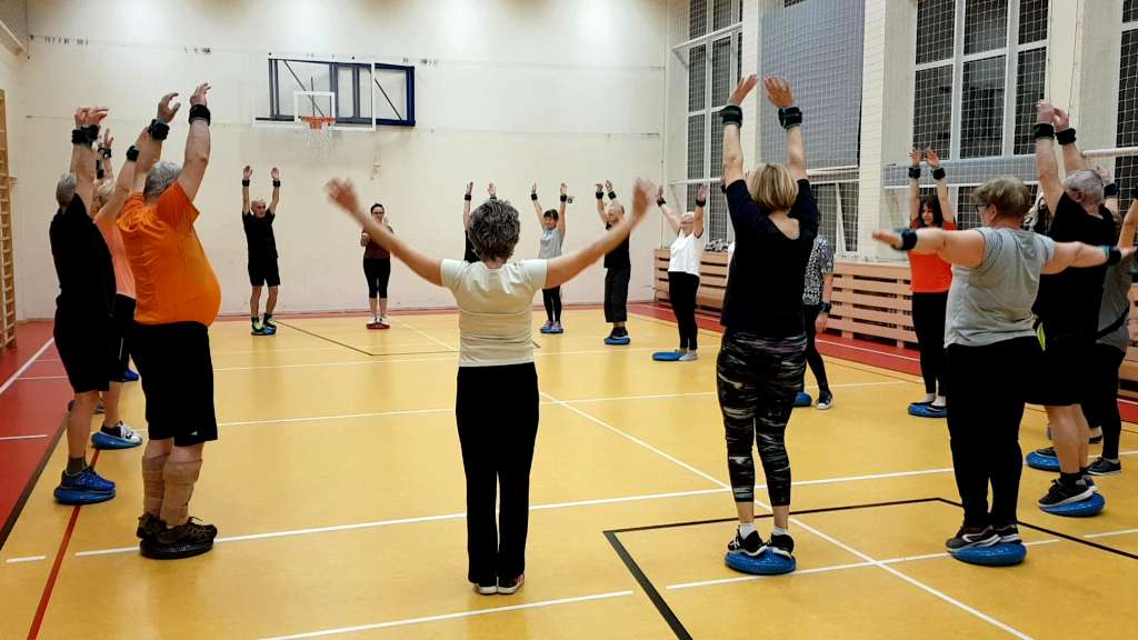 Ćwiczenia w TKKF Wierzbno. Grupa ćwicząca rozstawiona jest dookoła sali gimnastycznej. Uczestnicy stoją na tzw. beretach i wykonują ćwiczenia starają się utrzymać równowagę.