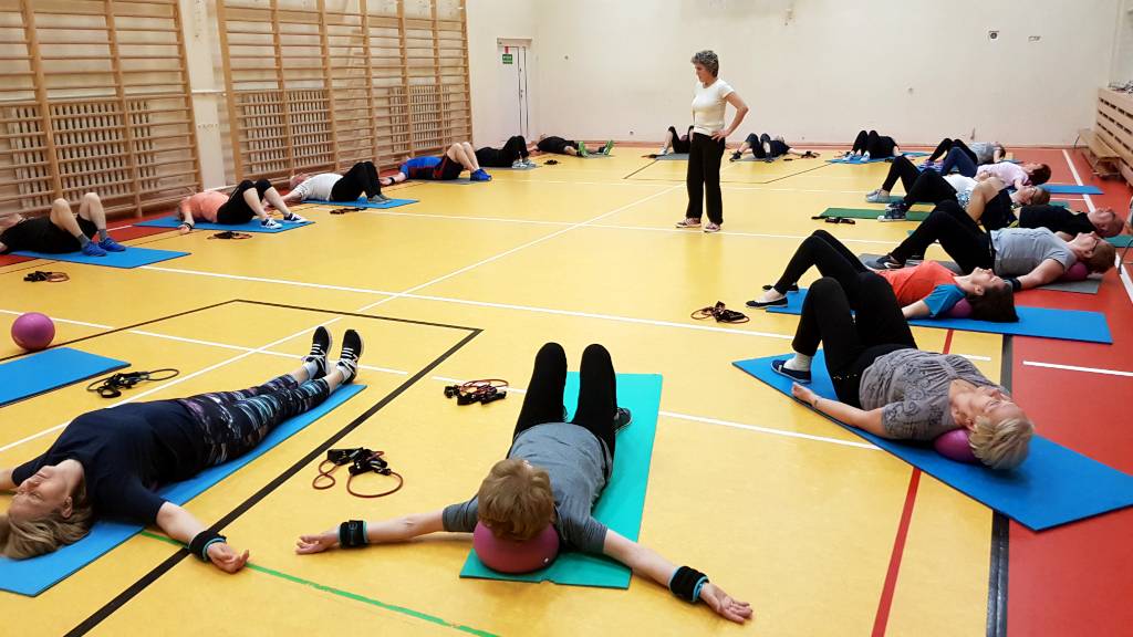 Gimnastyka dla seniorów - ćwiczenia relaksujące. Uczestnicy zajęć w pozycji leżącej, kolana zgięte, tęce na boku, szyja opiera się o piłkę. Ćwiczenia oddechowe.