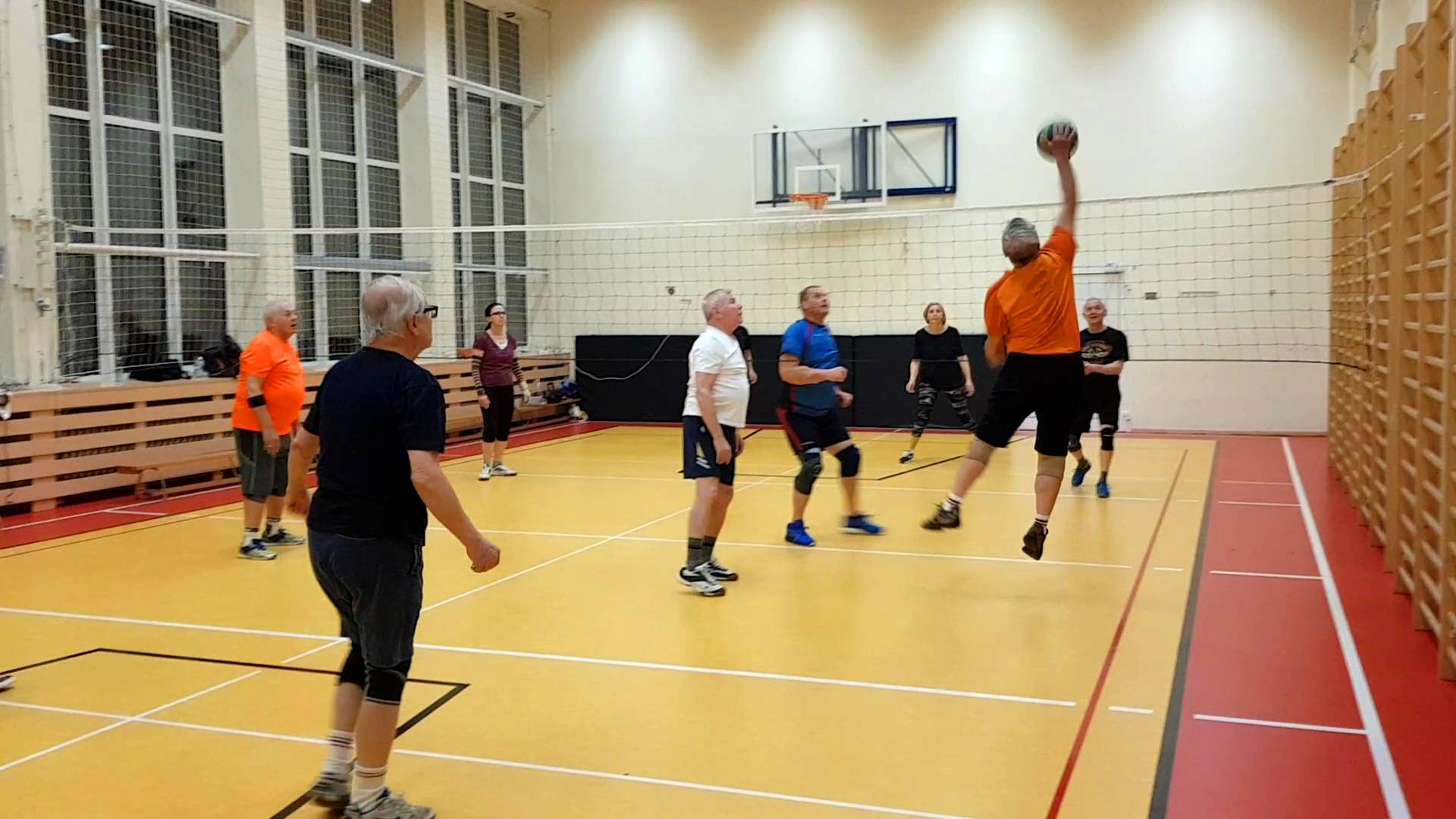 Siatkówka dla seniorów. Uczestnicy zajęć dla seniorów grają w siatkówkę. Zawodnik w pomarańczowej koszulce w wyskoku zbija.